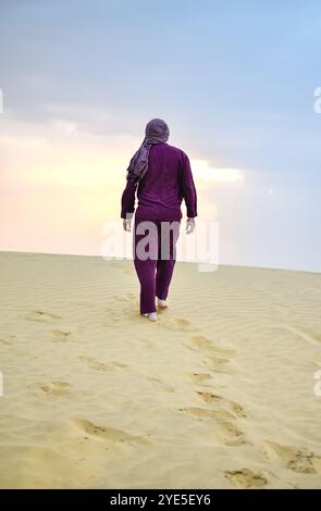 Un voyageur drapé dans une tenue violette marche délibérément à travers un désert serein, laissant des empreintes de pas dans le sable mou sous un ciel pâle à Jaisalmer, I. Banque D'Images