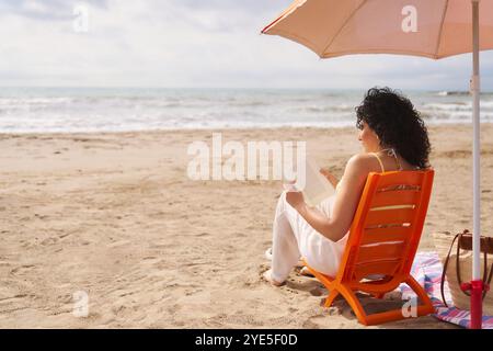 Une femme est assise sur une chaise de plage lisant un livre sous un parapluie. La scène est paisible et relaxante, avec le son des vagues en arrière-plan. T Banque D'Images