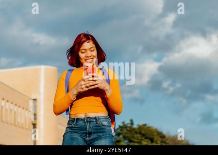 Une étudiante latine joyeuse aux cheveux roux saisissants est prise dans son smartphone, debout contre un campus en toile de fond sous un ciel bleu Banque D'Images