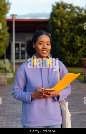 Une jeune étudiante joyeuse tenant un smartphone et un dossier, portant des écouteurs autour du cou, sur un campus au crépuscule Banque D'Images