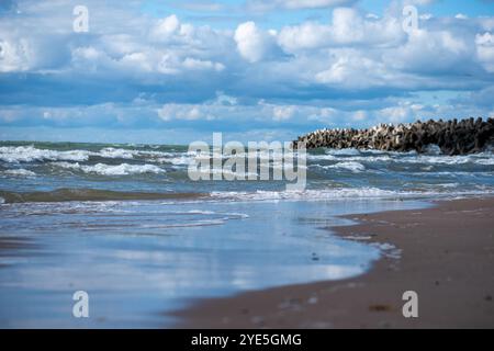 De douces vagues se glissent contre la plage de sable, reflétant le ciel nuageux tandis qu'une jetée rocheuse se jette dans l'eau, capturant la tranquillité d'une journée côtière. Banque D'Images