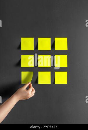Autocollants en papier jaune sur un mur noir gris foncé. Une main décolle une note adhésive. Banque D'Images