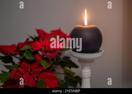 Une seule bougie scintille doucement sur un piédestal blanc, complété par des poinsettias rouges vifs à proximité, créant une atmosphère chaleureuse et accueillante pour le Banque D'Images