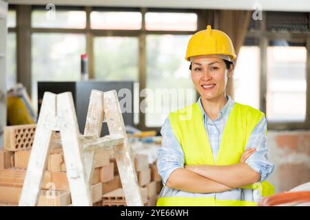 Portrait d'une femme de construction positive dans un gilet jaune et un casque de sécurité à côté d'un escabeau Banque D'Images
