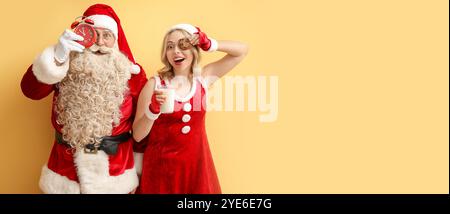 Père Noël drôle avec réveil et jeune femme en costume de Noël, avec verre de lait et biscuit sur fond jaune avec espace pour le texte Banque D'Images