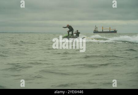 Un homme en combinaison noire monte une planche de surf en hydrofoil dans la mer tandis qu'un autre homme suit sur un jetski, au large de Sandbanks Beach, Poole, Dorset, Royaume-Uni. Banque D'Images