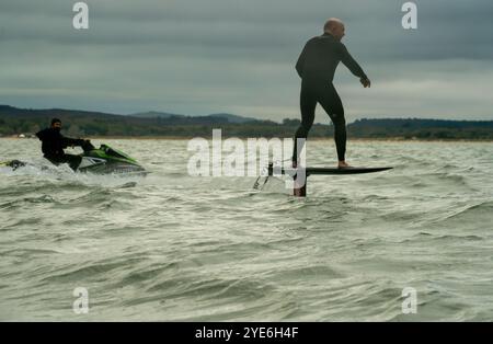 Un homme en combinaison noire monte une planche de surf en hydrofoil dans la mer tandis qu'un autre homme suit sur un jetski, au large de Sandbanks Beach, Poole, Dorset, Royaume-Uni. Banque D'Images