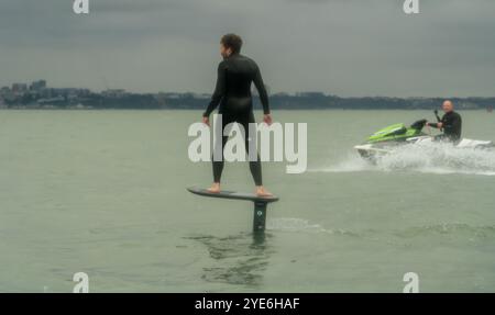 Un homme en combinaison noire monte une planche de surf en hydrofoil dans la mer tandis qu'un autre homme suit sur un jetski, au large de Sandbanks Beach, Poole, Dorset, Royaume-Uni. Studlan Banque D'Images