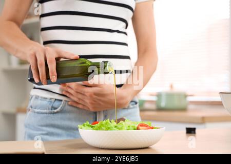 Jeune femme enceinte versant de l'huile dans une salade de légumes dans la cuisine, gros plan Banque D'Images