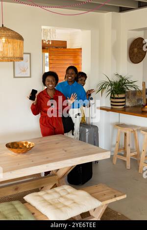 Amies afro-américaines arrivant avec des bagages, souriant et agitant dans un cadre confortable à la maison Banque D'Images