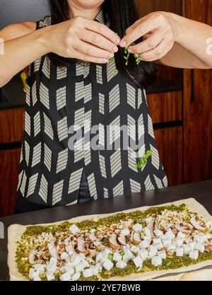 Une personne saupoudre du basilic fraîchement haché sur une pâte au pesto préparée, ajoutant une dernière garniture parfumée avant la cuisson. Banque D'Images