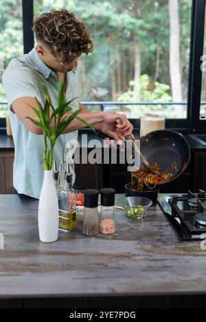 Homme cuisinant des nouilles dans une cuisine moderne, ajoutant des légumes du wok, à la maison Banque D'Images