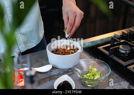 Homme garnissant les nouilles avec des herbes fraîches dans la cuisine moderne, préparant un repas délicieux, à la maison Banque D'Images