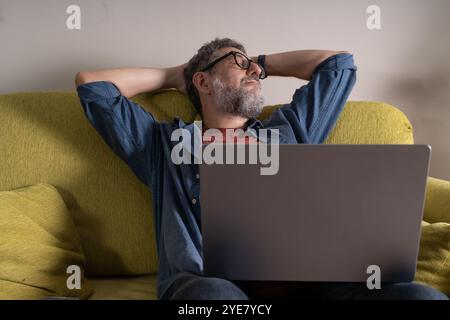 Professionnel d'âge moyen profitant d'un moment paisible pendant le travail à domicile. Portrait naturel de senior moderne prenant une pause détente sur le canapé jaune avec L. Banque D'Images