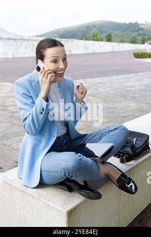 Heureuse femme d'affaires célébrant le succès tout en parlant sur le téléphone mobile et en tenant la tablette à l'extérieur Banque D'Images