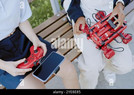 Prise de vue à angle élevé d'appareils électroniques dans des mains méconnaissables d'adolescents assis sur un banc en bois dans le parc Banque D'Images