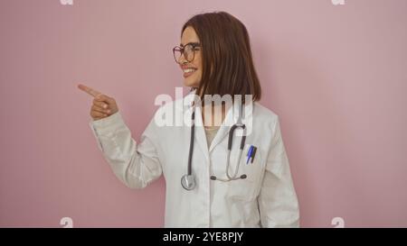 Une jeune femme hispanique attrayante médecin aux cheveux bruns sourit et pointe sur le côté tout en se tenant debout devant un mur rose isolé. Banque D'Images