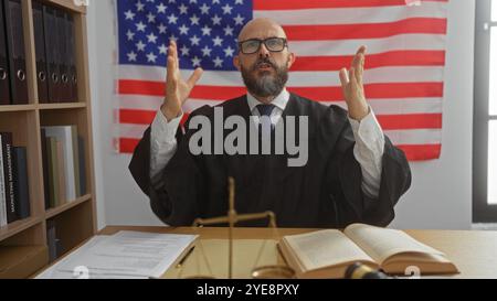 Bel homme hispanique portant une robe de juge faisant des gestes passionnés dans une salle d'audience américaine avec le drapeau des états-unis en arrière-plan. Banque D'Images