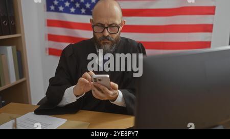 Homme hispanique dans la robe d'un juge utilisant un smartphone dans une salle d'audience américaine avec un drapeau américain en arrière-plan. Banque D'Images