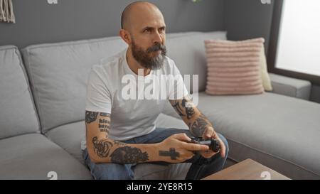 Homme tatoué jouant au jeu vidéo dans le salon montrant l'expression sérieuse sur le canapé, homme barbu chauve concentré à l'intérieur avec contrôleur et maison confortable int Banque D'Images