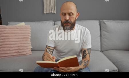 Bel homme hispanique avec une barbe assis à l'intérieur sur un canapé dans un salon écrivant dans un journal Banque D'Images