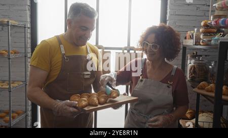 Homme et femme boulangers travaillant ensemble dans une boulangerie d'intérieur, arrangeant des pâtisseries fraîches sur un plateau en bois dans un intérieur de magasin lumineux avec des étagères de boulangerie Banque D'Images