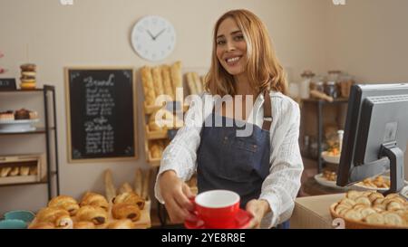 Femme hispanique travaillant à l'intérieur dans une boulangerie offrant des pâtisseries et du café avec le sourire, portant un tablier, avec du pain et des pâtisseries en arrière-plan Banque D'Images