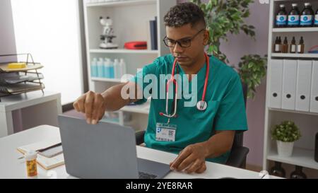 Médecin hispanique masculin portant des gommages et stéthoscope fonctionne sur un ordinateur portable dans un bureau de clinique avec des étagères et des fournitures médicales en arrière-plan Banque D'Images