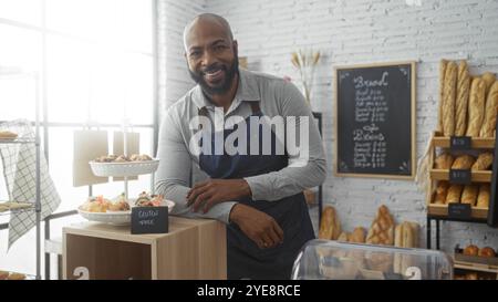 Beau jeune homme avec une barbe debout avec confiance dans une boulangerie portant un tablier, entouré de produits fraîchement cuits et un menu sur le mur Banque D'Images