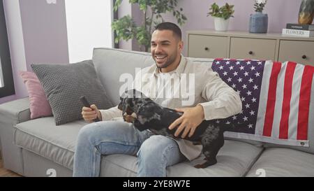 Un jeune homme hispanique tenant une télécommande de télévision est assis dans son salon avec son teckel, avec un drapeau américain drapé sur le canapé, entouré de confortable dec Banque D'Images