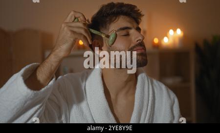 Bel homme dans une robe blanche utilisant un rouleau de jade sur son visage dans une salle de spa éclairée avec des bougies pour une ambiance relaxante Banque D'Images