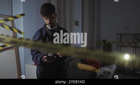 Un jeune homme hispanique avec une barbe examine des preuves sur une scène de crime intérieure faiblement éclairée, marquée par un ruban de mise en garde, dans un appartement moderne. Banque D'Images