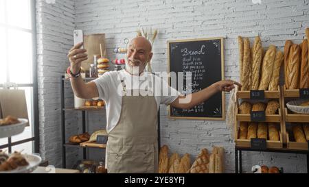 Homme senior tenant un téléphone sur un appel vidéo, présentant du pain dans une boulangerie confortable, souriant avec une barbe et portant un tablier sur fond de BA Banque D'Images