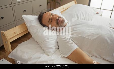 Homme hispanique d'âge moyen reposant dans un lit dans un intérieur de chambre serein avec draps blancs et meubles en bois. Banque D'Images