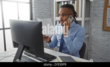 Jeune homme avec des tresses travaillant dans un bureau, parlant au téléphone tout en examinant des documents à un bureau, mettant en valeur un cadre intérieur professionnel avec M. Banque D'Images