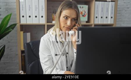 Une jeune femme blonde caucasienne attrayante dans une clinique faisant un appel à partir d'un téléphone de bureau tout en travaillant sur un ordinateur dans un bureau empilé avec bi Banque D'Images