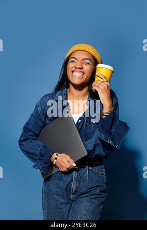 Une jeune femme joyeuse dans des vêtements chauds d'automne tient une tasse à café et un ordinateur portable, exsudant la joie. Banque D'Images