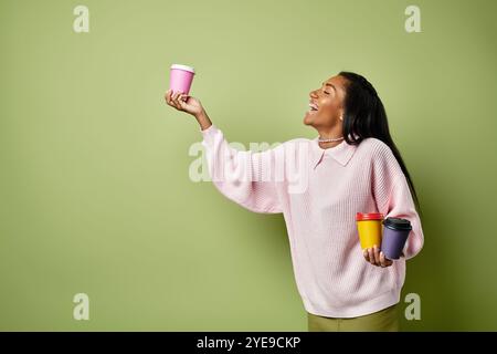 Une jeune femme sourit largement, équilibrant des tasses de café colorées sur un fond vert. Banque D'Images