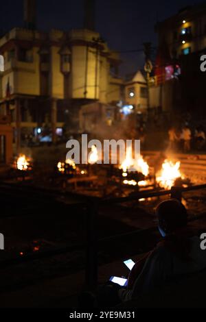 Les écrans de téléphones intelligents brillent au premier plan avec des corps brûlants au Manikarnika Ghat, un sanctuaire hindou et un terrain de crémation le long du Gange en... Banque D'Images