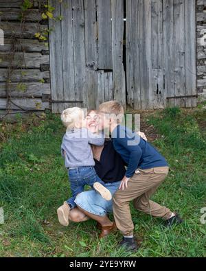 Portrait d'une mère assise sur l'herbe devant un bâtiment en bois avec ses deux fils qui l'embrassent et l'embrassent, canton de Beckwith à Carleton PL... Banque D'Images