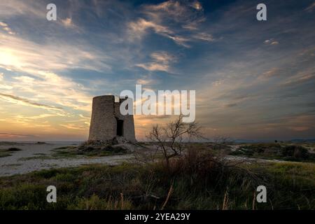 Un vieux moulin à vent en pierre sur une plage au coucher du soleil avec la lumière du soleil magnifique et les nuages du ciel. Banque D'Images