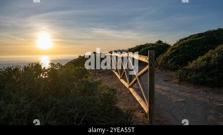 Vue panoramique sur l'océan et la plage de Benagil au coucher du soleil le long de la côte du Portugal, avec une barrière de rail en bois le long d'un sentier de sable à un point d'observation Banque D'Images