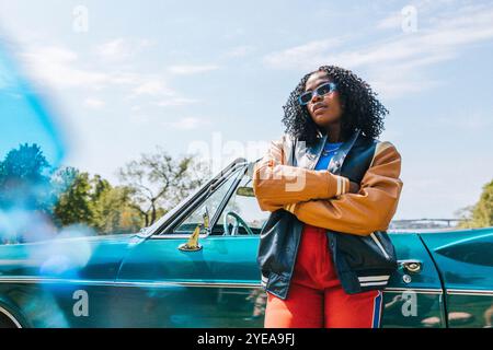 Femme aux cheveux bouclés confiants avec les bras croisés debout près de la voiture sous le ciel Banque D'Images