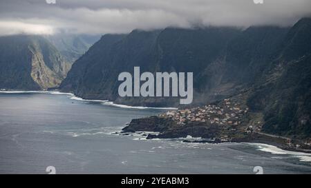 Vue depuis Eira da Achada point de vue des falaises escarpées le long de la côte sous les nuages d'orage et les maisons à Ribeira da Ja sur l'île de Madère... Banque D'Images