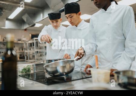 Stagiaires adolescents masculins préparant la nourriture par une collègue féminine tenant une casserole dans une cuisine commerciale Banque D'Images