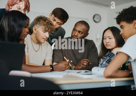 Adolescente discutant avec un enseignant masculin tout en étant assise avec des camarades de classe au bureau Banque D'Images