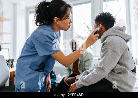 Expert médical féminin vérifiant l'oreille du garçon avec un thermomètre dans la salle d'examen Banque D'Images