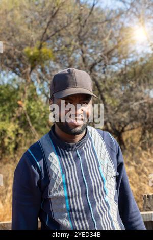 homme africain souriant avec une barbe portant une casquette, coucher de soleil du village, buisson en arrière-plan Banque D'Images