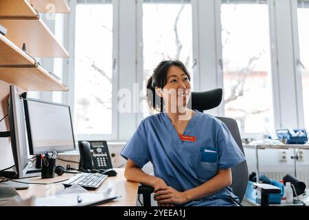 Médecin féminin souriant regardant loin tout en étant assis sur la chaise près de la table dans la salle d'examen Banque D'Images