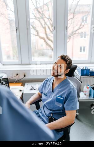 Vue en angle élevé d'un expert médical masculin portant des gommages bleus assis sur une chaise et parlant avec un collègue à la clinique Banque D'Images